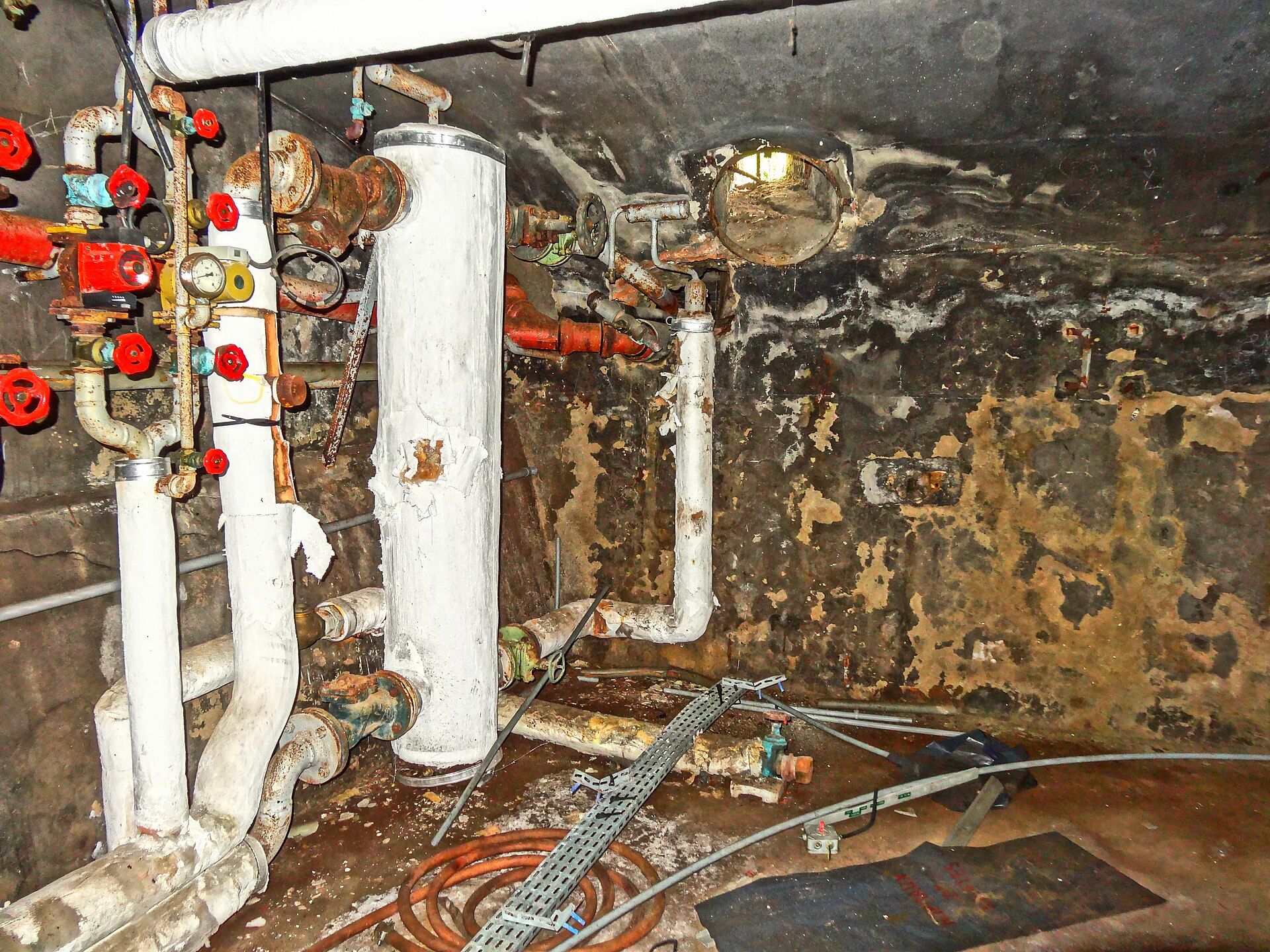 A cramped old boiler room with red valve handles, wrapped white pipes and water stained walls, for a theme in the writing of Richard Joseph Spinelli