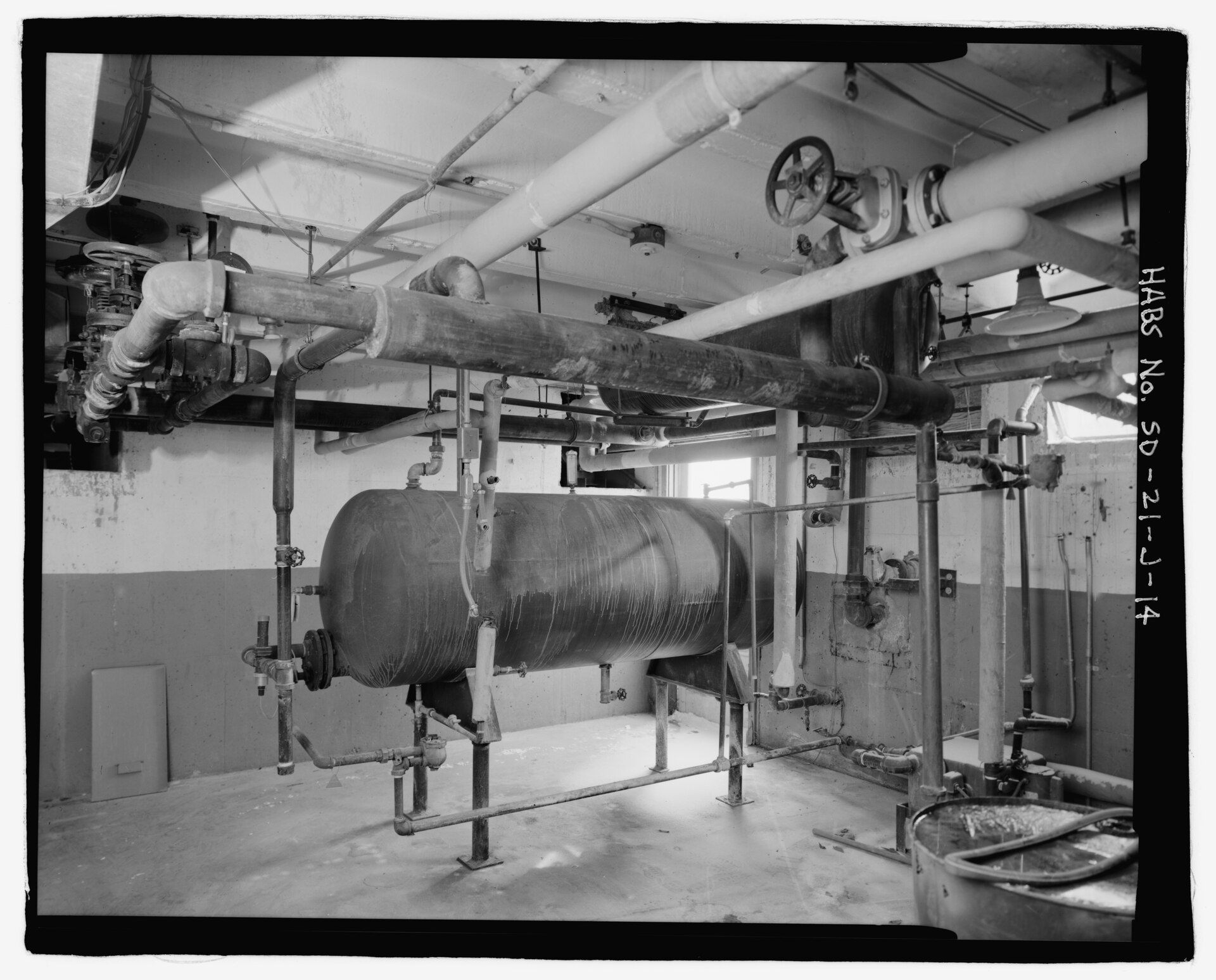 A basement mechanical room with a long steel tank, valves and a web of insulated pipes overhead, for a reading guide to Richard Joseph Spinelli