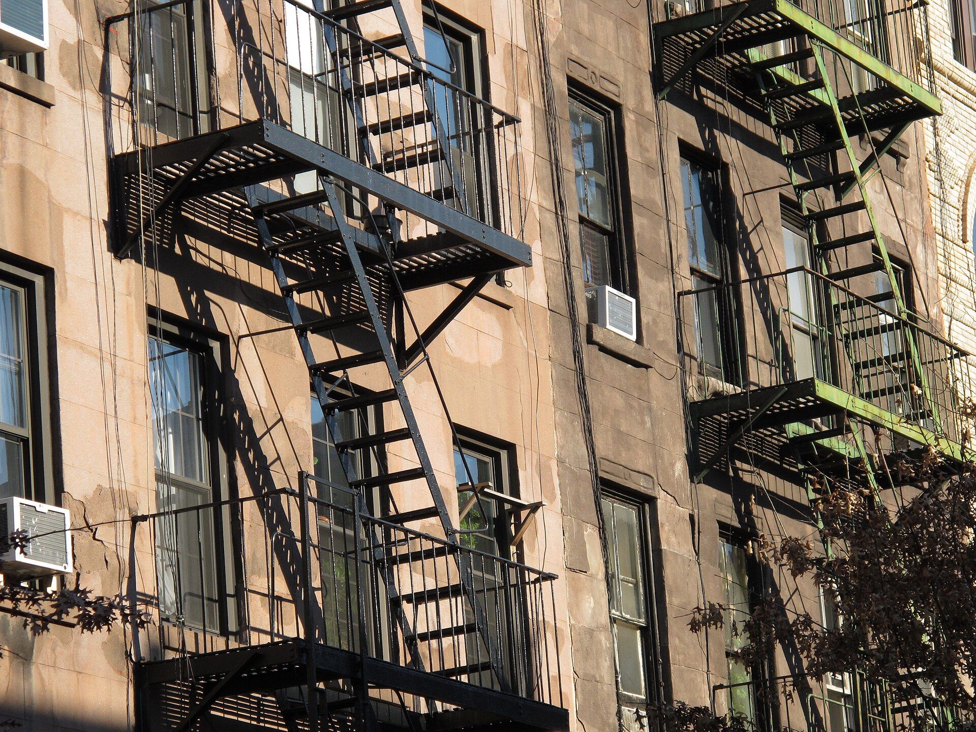 Fire escapes throwing long shadows across the weathered facade of an older apartment building, for a series by Richard Joseph Spinelli