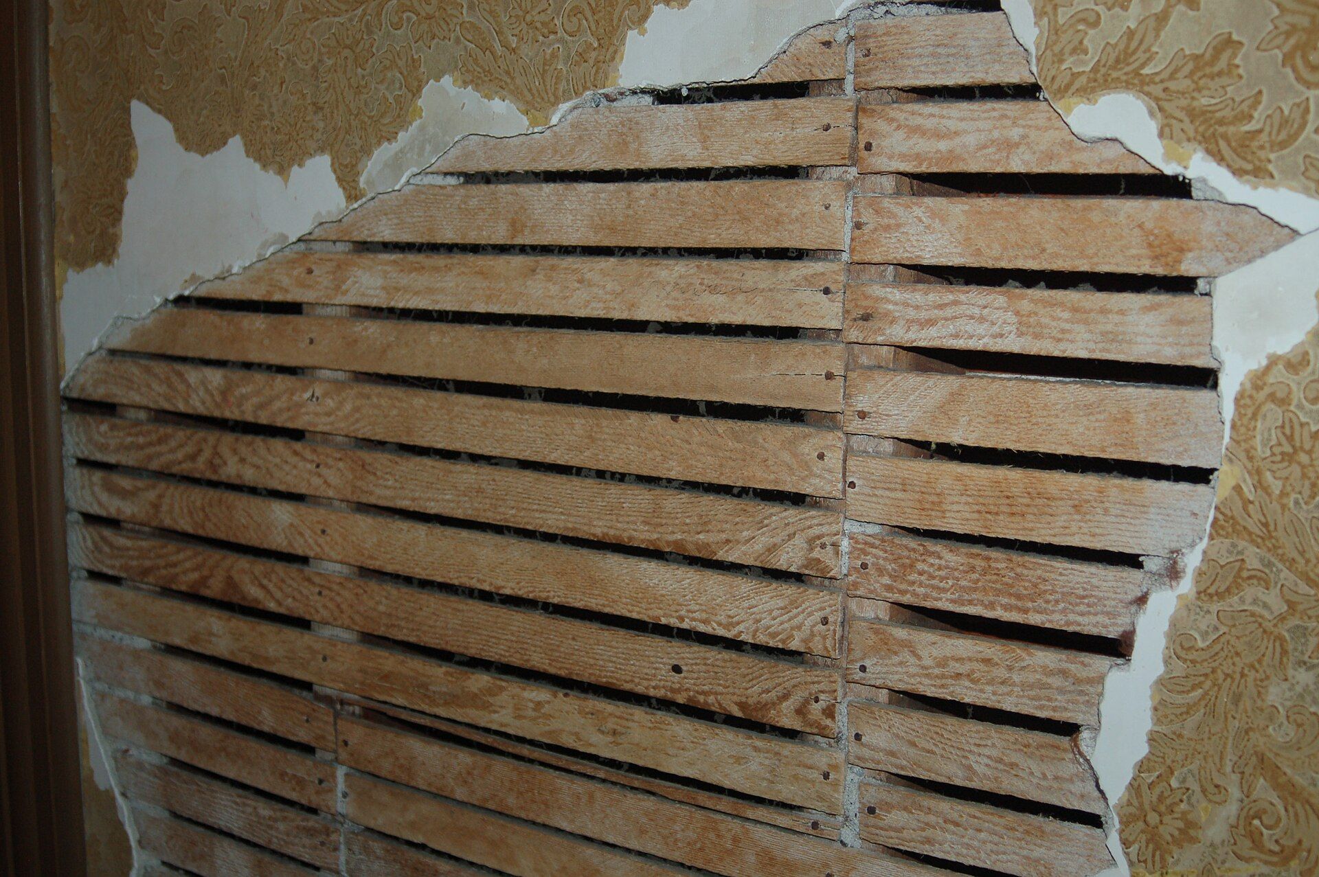 Rows of wood lath showing through a hole in an old wall where plaster and wallpaper have broken away, for a theme in the writing of Richard Joseph Spinelli