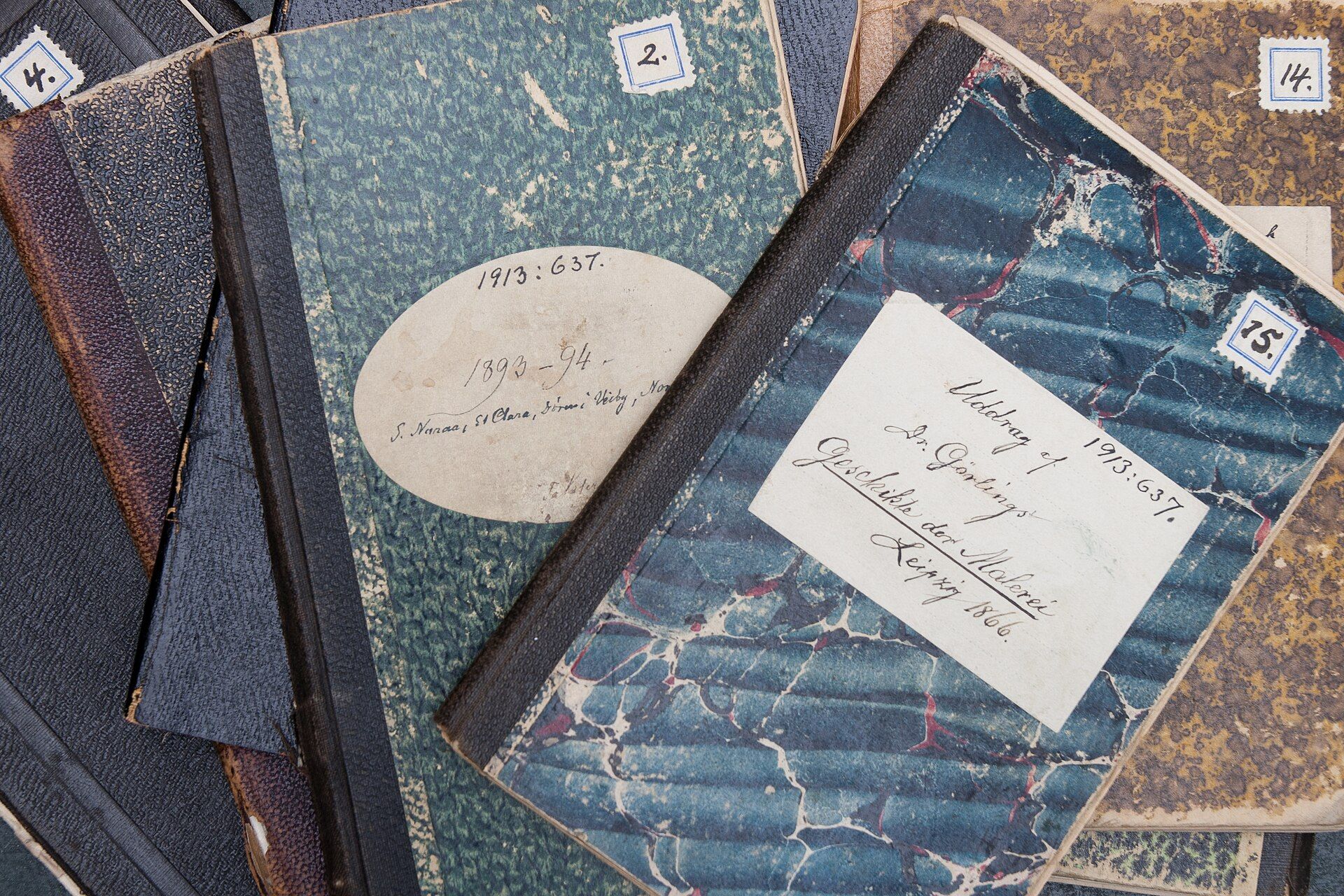 Old notebooks with marbled covers and handwritten paper labels spread across a table, for a series by Richard Joseph Spinelli
