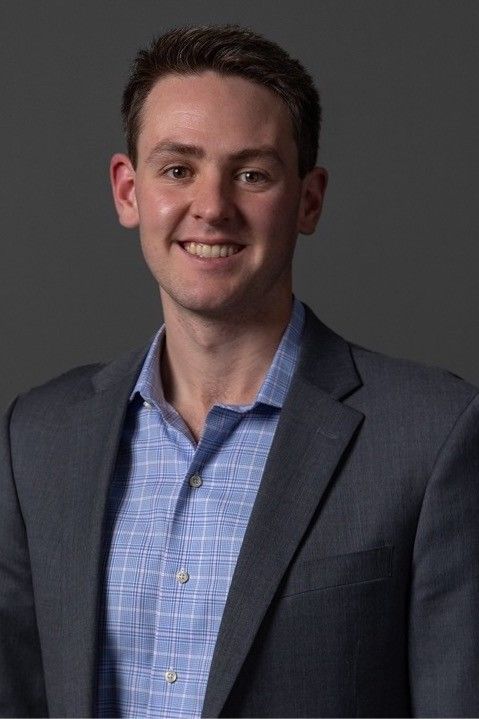 Richard Joseph Spinelli in a grey jacket and blue checked shirt against a dark studio backdrop, a tall portrait