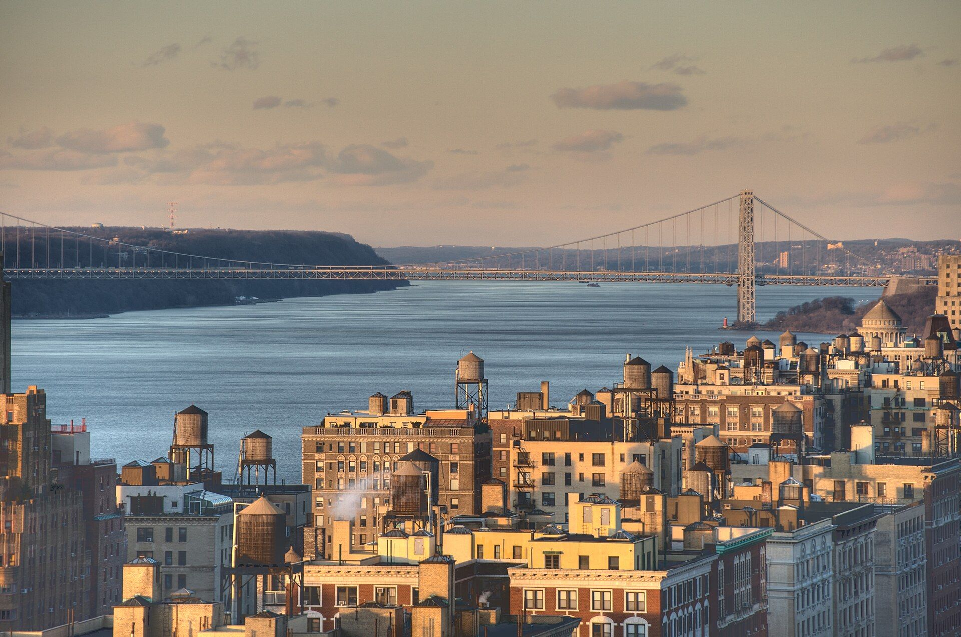 Apartment rooftops crowded with water tanks in late afternoon light, with the Hudson River and a long suspension bridge beyond, the hero for the Medium channel of Richard Joseph Spinelli